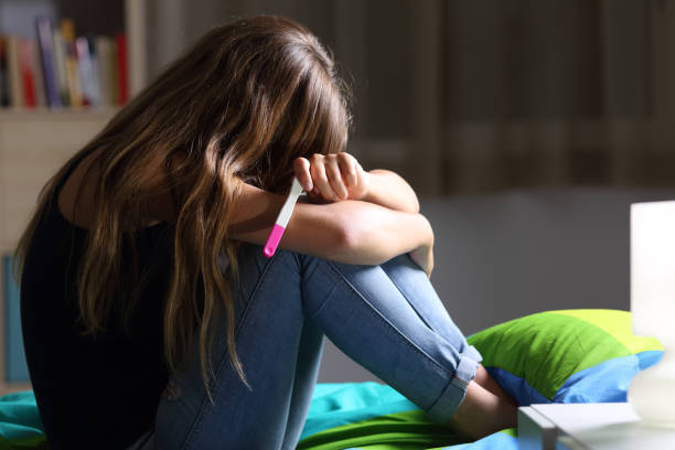 Sad pregnant teen after pregnancy test Side view of a sad pregnant teen sitting on her bed after checking a pregnancy test with a dark light in the background unwanted pregnancy stock pictures, royalty-free photos & images