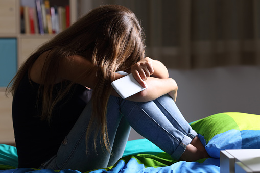 Single sad teen holding a mobile phone lamenting sitting on the bed in her bedroom with a dark light in the background