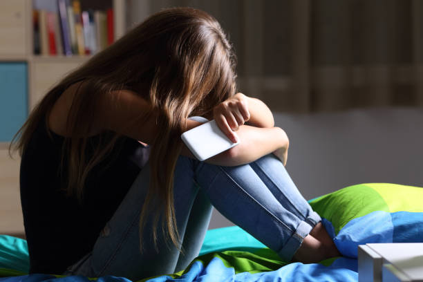 Sad teen with a phone in her bedroom Single sad teen holding a mobile phone lamenting sitting on the bed in her bedroom with a dark light in the background victim photos stock pictures, royalty-free photos & images