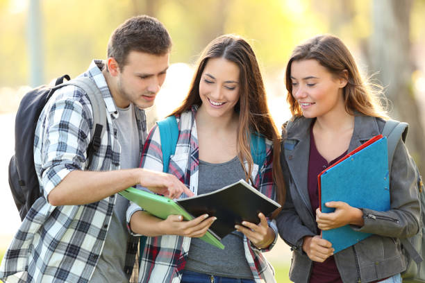 Three students learning reading notebook Front view of three students learning reading a notebook and commenting in the street teenager adolescence campus group of people stock pictures, royalty-free photos & images