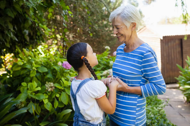 femme senior souriante, main dans la main de la petite-fille en se tenant debout contre les plantes - generation gap multi generation family vertical holding hands photos et images de collection