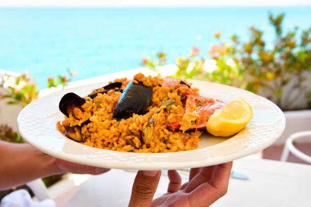 closeup of a young man bringing a plate of typical spanish paella, with seafood, to a table set for lunch, with the sea in the background