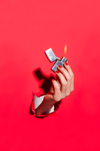 Vertical studio shot of hand through hole in red cardboard sheet holding flaming lighter.