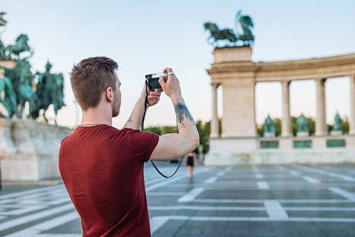 Young man, 20 years ld, discovering Eastern European cities and taking photos of famous landmarks. Young man is taking photos of famous Hero's Square in Budapes, Hungary and sharing them to social media.