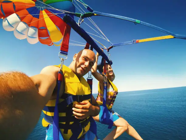 Selfie of people enjoying parasailing over sea on a summer day during their vacation. They are being pulled by a speedboat.