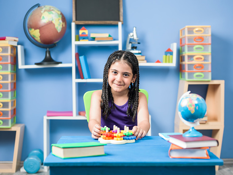 Portrait of 7 years old schoolgirl wearing a purple dress sitting on chair and playing with toy gear puzzle on desk.She has long hair  braids.Books,globe,blackboard and other education equipments are seen on bookshelf on the background.The background is defocused.Shot indoor.Horizontal framing.