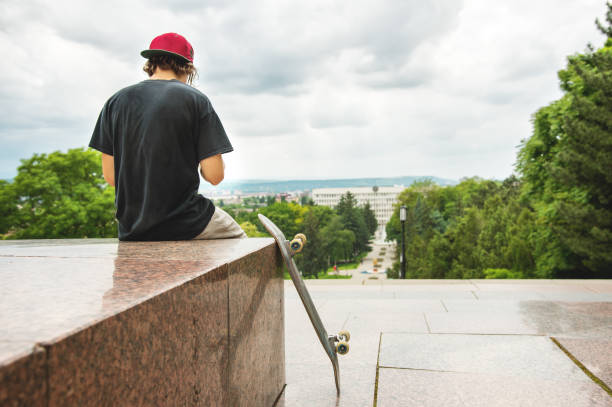 The long-haired skater in a cap and T-shirt sits with his back to the camera and thinks next to the skateboard against the background of the landscape landscape.