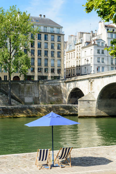 istock Two deck chairs in the sun under a parasol in front of the river Seine. 820774554