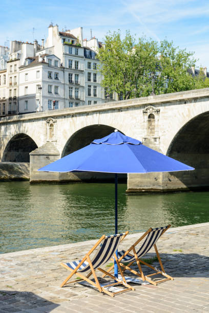 istock Two deck chairs in the sun under a parasol in front of the river Seine. 820775924