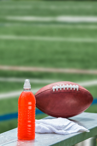 Selective focus on an ice cold plastic bottle of orange flavored sports drinking sitting on an aluminum bench with a leather American football and white towel with a football playing field in the background . It is important to keep hydrated while playing sports or other activities on a hot day.