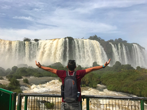 Young backpacker male tourist open his arms in Iguazu National Park, Brazil carrying a backpack in a nice summer day with a beautiful waterfalls on back.