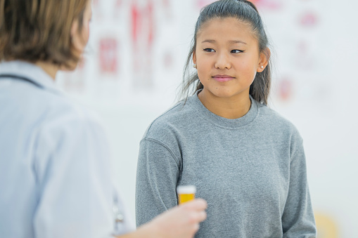 An Asian girl is indoors in a physiotherapy clinic. She is wearing casual clothing. A female doctor wearing a lab coat is giving her medication for her injury.