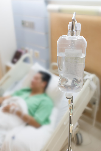 Blurred image of male patient lying on hospital bed, with saline solution bag as foreground.