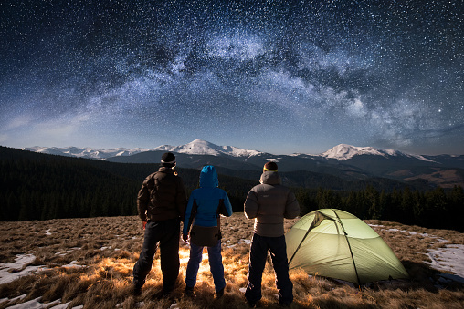 Rear view couple hikers holding hands and their friend standing near green tent in the camping under beautiful night sky full of stars and milky way. On the background snow-covered mountains
