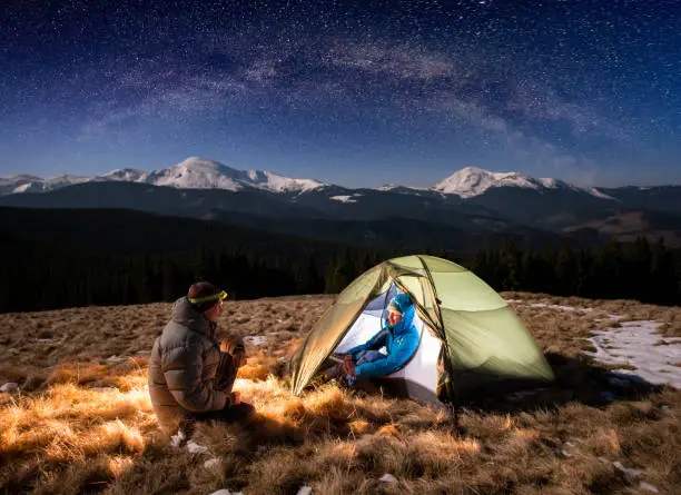 Photo of Young couple tourists having a rest in the camping at night, sitting near campfire and green tent under beautiful night sky full of stars and milky way. On the background snow-covered mountains