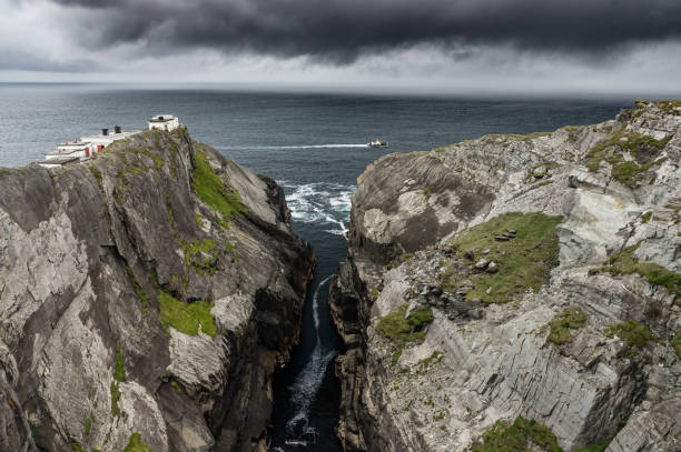 Ferry crossing rocky headland of Mizen Head, County Cork, Ireland, with dark storm clouds approaching.