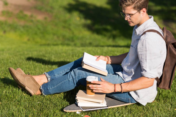 istock handsome young man in spectacles reading book and drinking coffee while sitting on skateboard in park 820860466