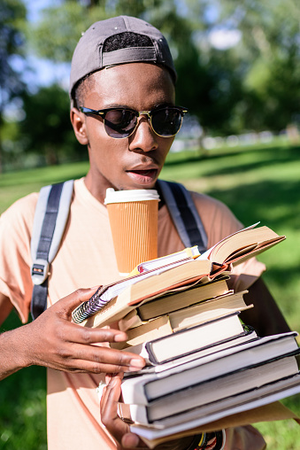 young african american man in cap and sunglasses holding pile of book with paper cup
