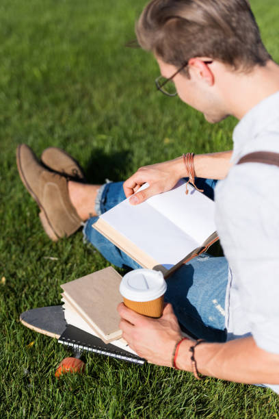 istock high angle view of young man in eyeglasses and backpack reading book while sitting on skateboard in park 820863302