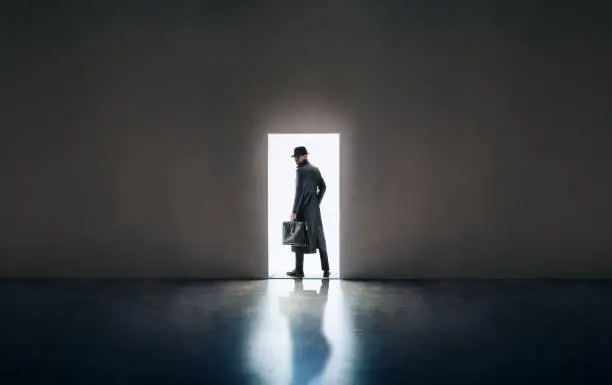 Man silhouette in hat and raincoat standing in the light of opening door in dark room