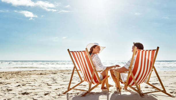 Soaking up some good times together Shot of a mature couple relaxing together on deck chairs at the beach deckchair stock pictures, royalty-free photos & images