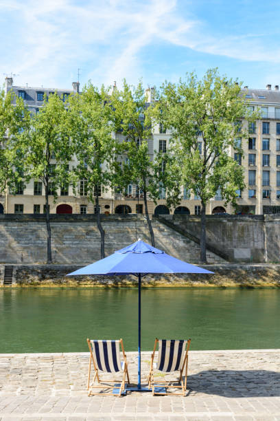 istock Two deck chairs in the sun under a parasol in front of the river Seine. 820874658