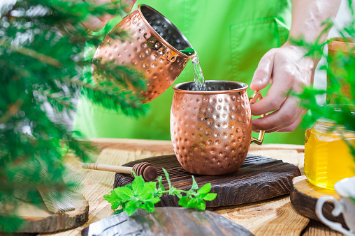 Person wearing green dress making cold drink in copper mug. Honey and herbs on wooden table. Midsection view