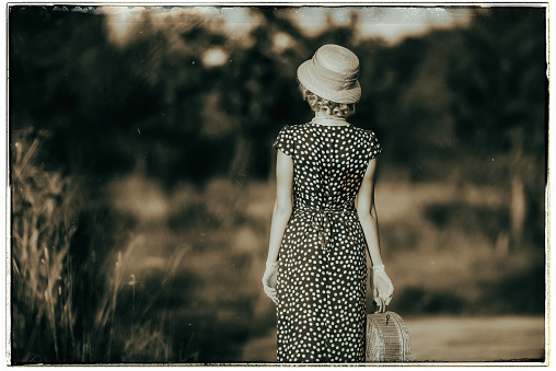 Classic black and white photo of vintage 1920s summer fashion woman with dress and straw hat standing with handbag in rural landscape. Rear view.