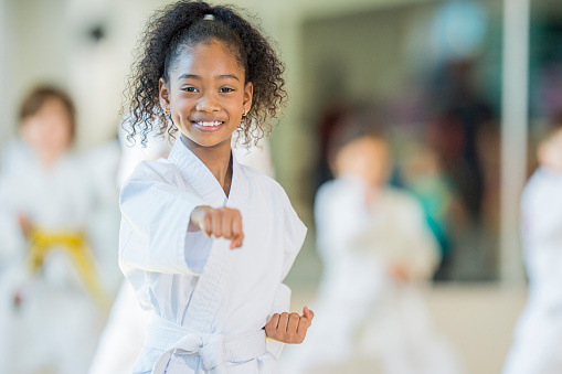 A multi-ethnic group of children are indoors at a Taekwondo academy. They are wearing martial arts clothing. A girl of African descent is punching while smiling at the camera.