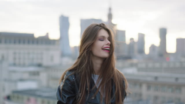 Pretty woman with long straight brown hair looking at camera. Big city in background.