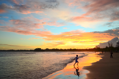Sunset in the tropics on the ocean coast. The child is jumping on the beach. Bright colors of the sky are reflected in the water and sand