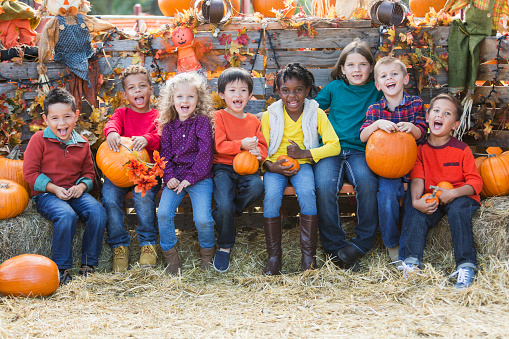A group of eight multi-ethnic children sitting in a row surrounded by pumpkins, scarecrows and hay. They are at a fall festival celebrating autumn.