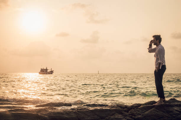 A striking view of a towering limestone karst formation at Railay Beach, Krabi, Thailand, silhouetted against the soft glow of the setting sun. People wade through the shallow water, adding a sense of scale and tranquility to this tropical coastal scene. The dramatic rock formation and calm waves reflect the untouched beauty of Thailand's Andaman Sea coastline.