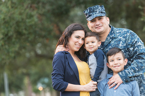 An Hispanic man in the US military, wearing camouflage clothing, standing with his sons, 2 and 8 years old, and wife outdoors in the park.