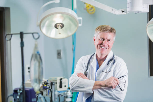 Male doctor wearing white lab coat Portrait of a male doctor in his 50s wearing a white lab coat, standing in an operating room with surgical lights and medical equipment, looking at the camera. surgical light stock pictures, royalty-free photos & images