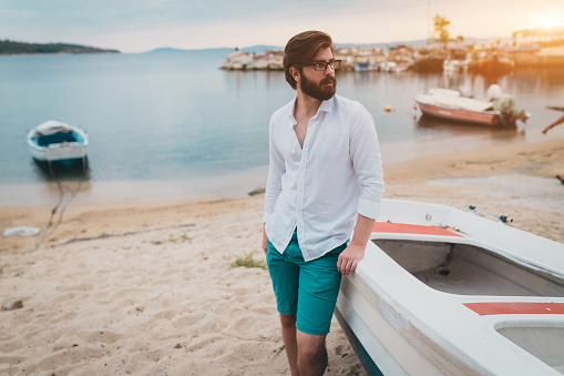 Young handsome bearded man standing by the boat at the beach