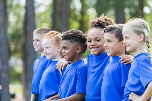 A group of six multi-ethnic children 6-7 years old playing soccer at summer camp. They are standing in a row smiling in their uniforms of royal blue shirts in the park. One African-American girl is looking at the camera.