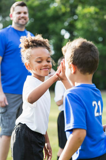 Multi-ethnic group of children, 6-7 years old playing soccer. An African-American girl is giving a high-five to a boy from the other team. The coach of the blue team is holding a soccer ball.