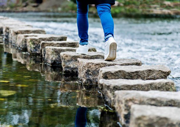 femme traversant les pierres de progression sur une rivière - stepping stone stone stepping footpath photos et images de collection