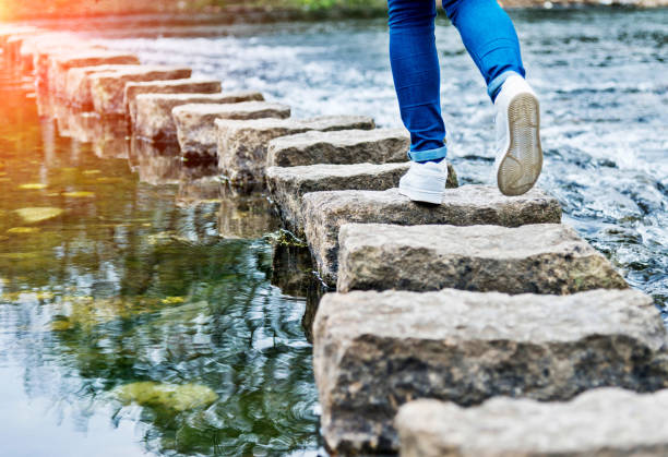 femme traversant les pierres de progression sur une rivière - stepping stone stone stepping footpath photos et images de collection