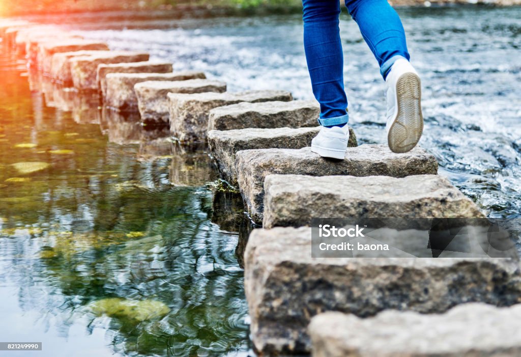 Woman crossing stepping stones on a river The Next Step Stock Photo