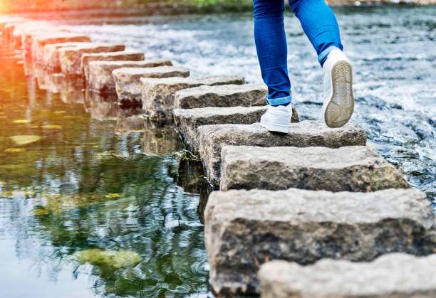 woman crossing stepping stones on a river - stone walkway imagens e fotografias de stock