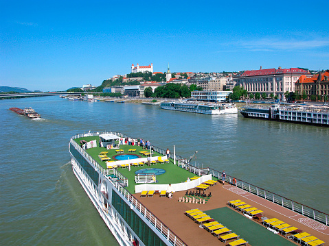 People travel on Danube river on large cruise ship. They looking at panorana of Bratislava city. On hill above the city is Bratislava castle. Close to castle is St. Martin cathedral. On cruise ship has round swimming pool, game area for chess, sunbeds on deck.