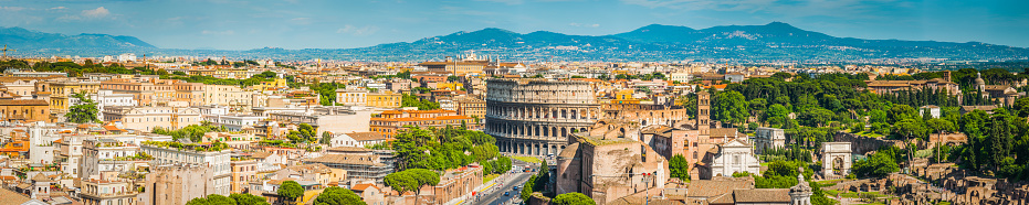 Panoramic view across the landmarks of Rome, Italy, from the Via dei Fori Imperiali and iconic arches of the Coliseum, the ancient ruins of the Roman Forum, Esquilino, Aventino and Palatine Hills.