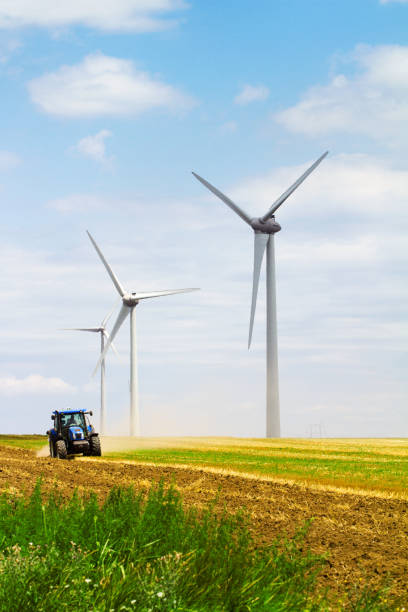 row of eolian wind turbine on a meadow with tractor ploughing the land in the background on a sunny day blue sky with clouds