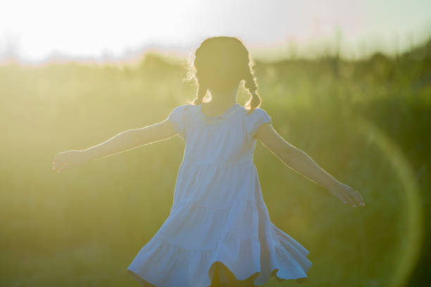 An Ethnic girl is outdoors in a field on a sunny day. She is wearing a blue dress. She is standing with her arms out while the sun sets.