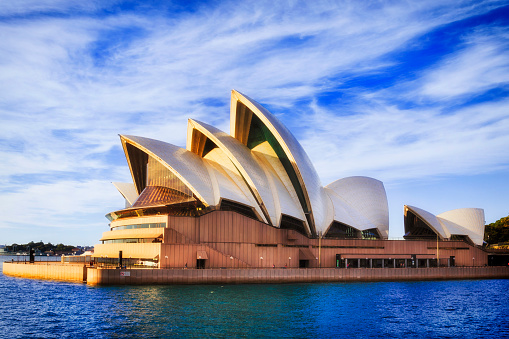 Sydney: Sydney opera house theatre building rising from Sydney harbour waters on a sunny bright day against blue sky.