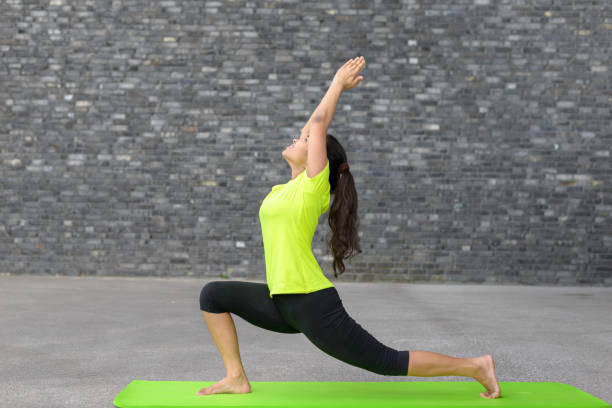 Fit athletic young woman doing yoga exercises Fit athletic young woman doing yoga exercises stretching and arching her back with raised arms in a profile view on a mat in front of the wall of an urban building with copy space warrior position stock pictures, royalty-free photos & images