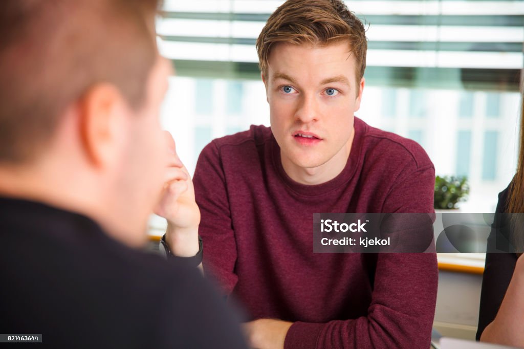 Businessman Looking At Colleague While Communicating In Office Confident young businessman looking at male colleague while communicating in office Discussion Stock Photo