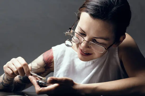 Colour portrait of a female watchmaker or watch repairer at work. In her hand you can see a watch that he trying to repair, she is wearing specialist magnifying glass and on her workbench are various watchmaking tools. Horizontal format  with the focus on the watch in her face.